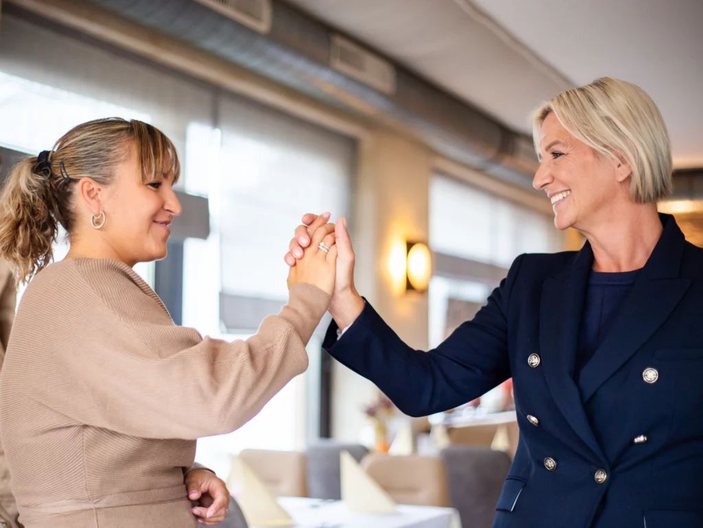 Claudia Angela Ruttnig gibt einer Dame einen freundlichen Handschlag. Im Hintergrund ist ein Gastraum zu sehen.
